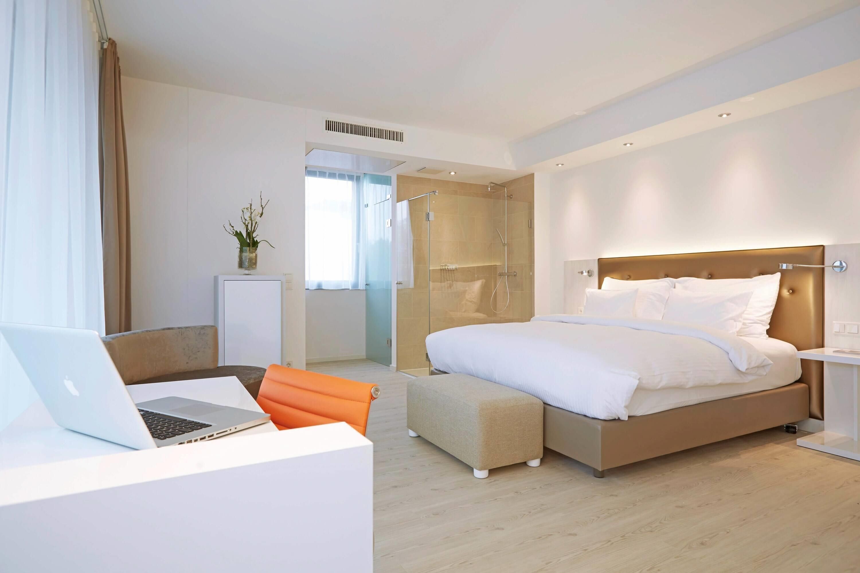 A modern hotel room with a bed, a desk with a laptop, a shower area, and a small seating area.
