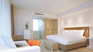 A modern hotel room with a bed, a desk with a laptop, a shower area, and a small seating area.