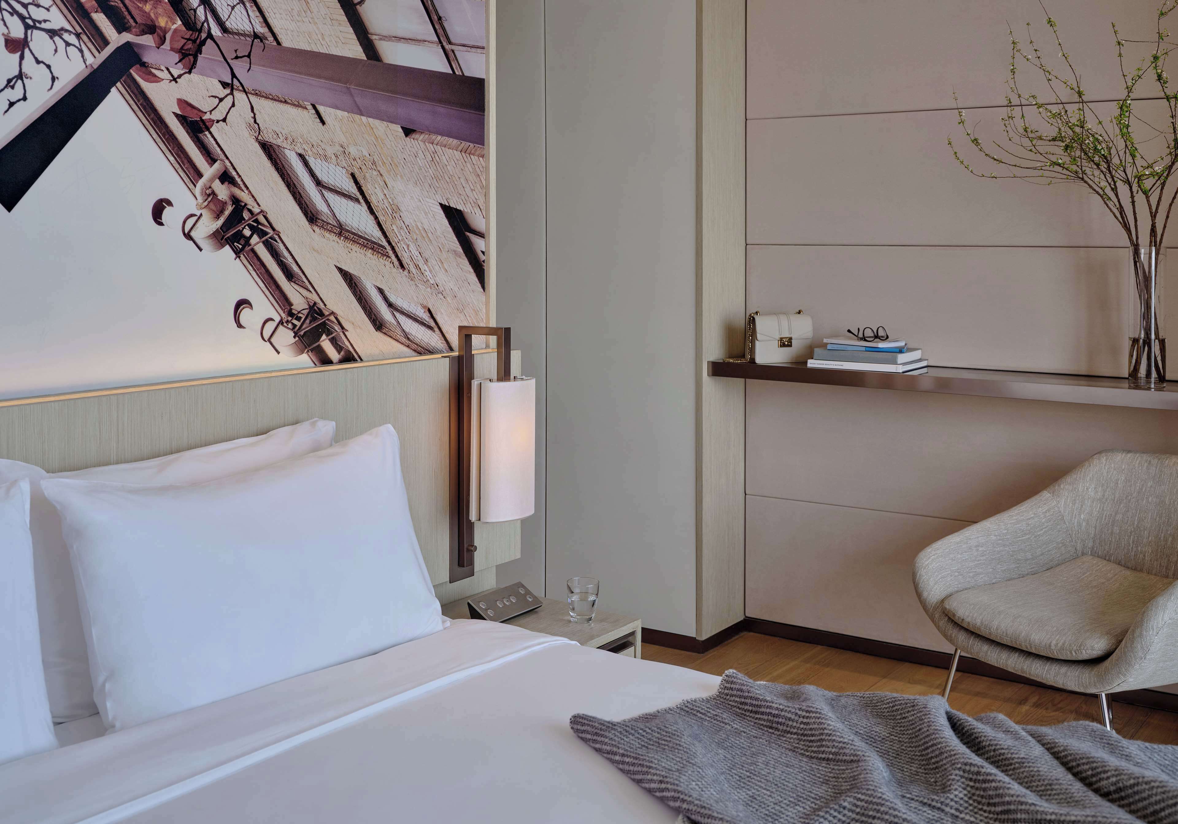 A hotel room with a bed, a chair, a wall-mounted shelf with books and a vase, and a painting on the wall.