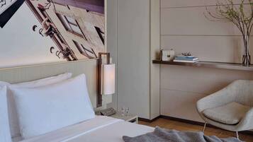 A hotel room with a bed, a chair, a wall-mounted shelf with books and a vase, and a painting on the wall.