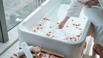Una persona en una bañera con flores flotando en el agua, una copa de sales de baño y una bandeja con flores sobre un banco de madera.