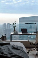 A modern hotel room with a large window, a desk with a laptop, a vase with a plant, a chair, and a bed.