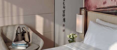 A hotel room with a bed, a chair with a handbag, a vase with flowers, and a wall with Chinese characters.