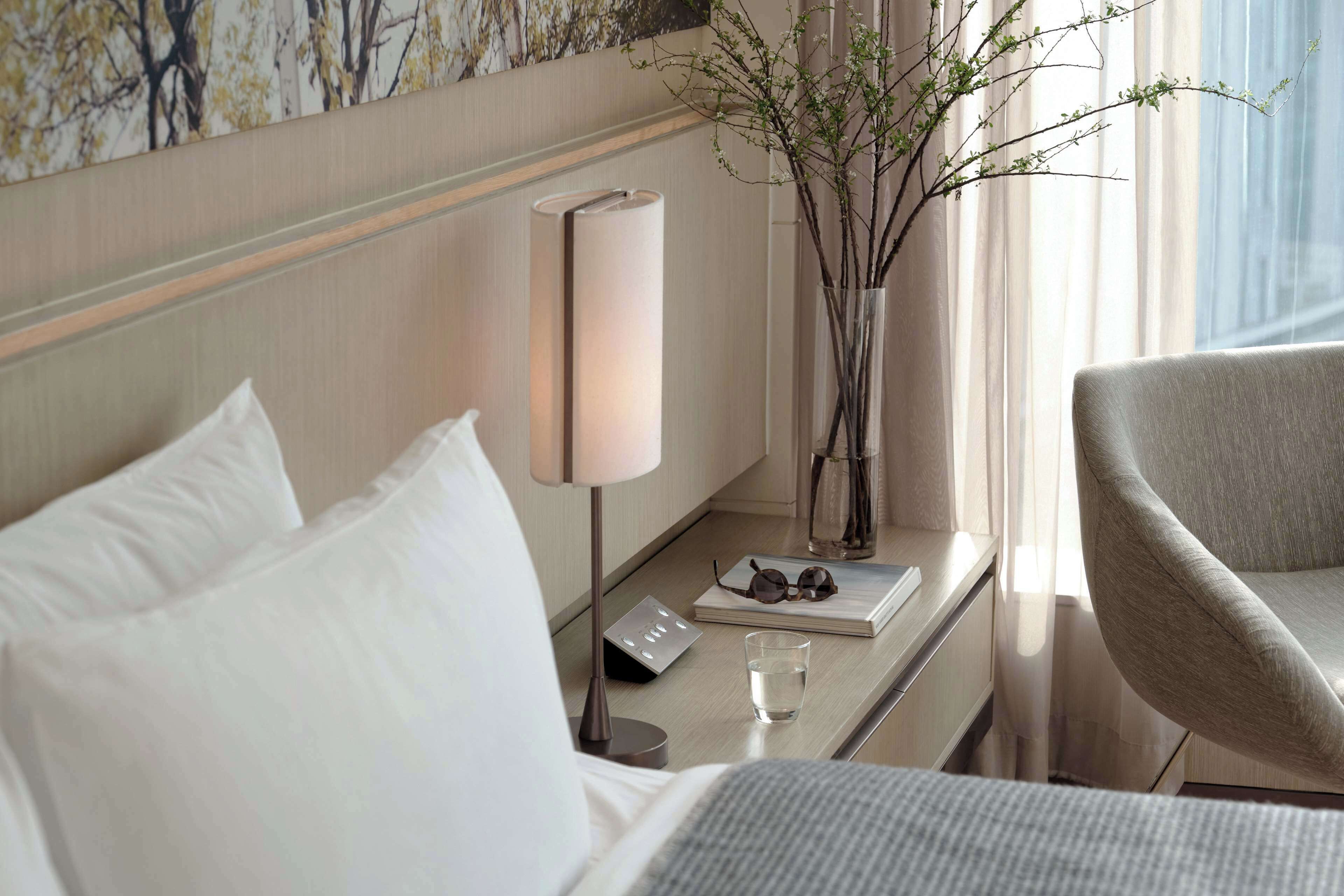 A hotel room with a bed, a bedside table with a lamp, a vase with branches, a chair, and a window with curtains.