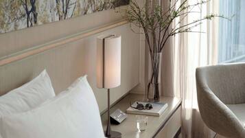 A hotel room with a bed, a bedside table with a lamp, a vase with branches, a chair, and a window with curtains.