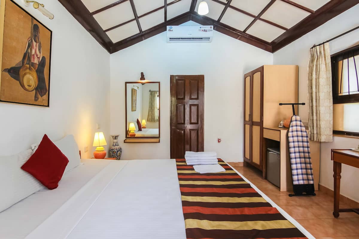 Habitación de hotel con cama, almohada roja, alfombra a rayas, espejo, puerta, tabla de planchar y ventana con cortinas.