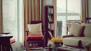 A cozy living room with a sofa, a chair, a bookshelf, and a view of the outdoors through large windows. - Villa Paradiso (Dharamshala)