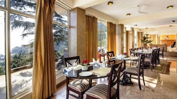 A dining area with large windows, wooden chairs, and a table set for a meal. - Villa Paradiso (Dharamshala)