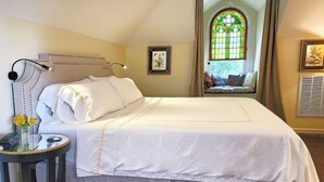 Cama bem arrumada com colcha branca e cabeceira decorativa. Uma pequena mesa com um vaso de flores está ao lado da cama.