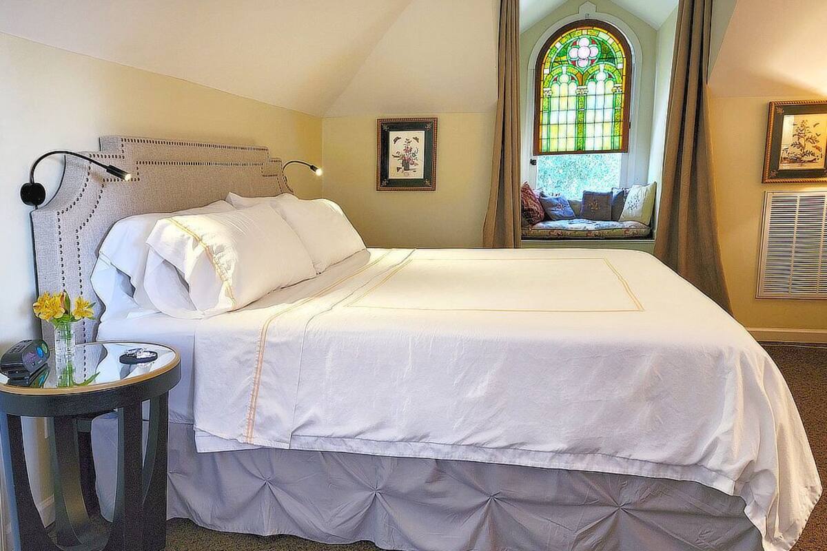 Un lit bien fait, recouvert d’une housse de couette blanche et agrémenté d’une tête de lit décorative. Une petite table avec un vase de fleurs se trouve à côté du lit.