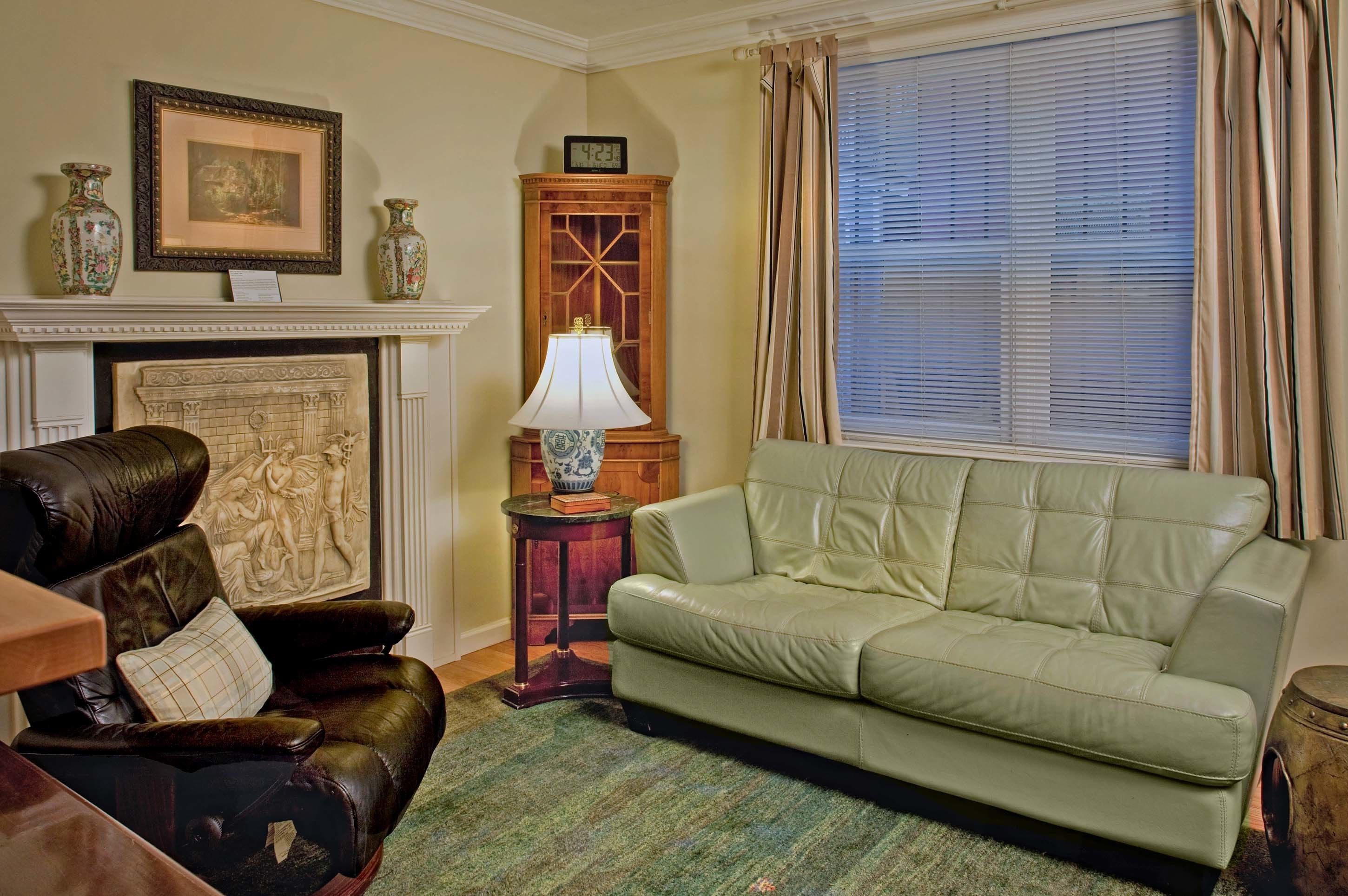 A living room with a green sofa, a leather armchair, a fireplace, a clock, and a window with curtains.