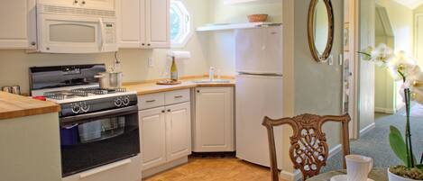 A kitchen with white cabinets, a microwave, stove, and refrigerator. There's a dining table with a chair and a potted plant.