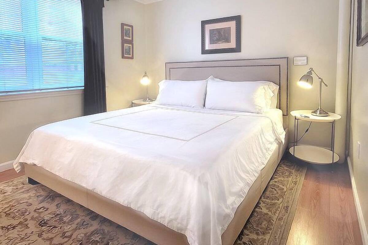 Un lit bien fait, avec du linge de lit blanc, se trouve dans une pièce dotée d’un plancher en bois et d’un tapis à motifs. Deux tables de chevet avec des lampes sont disposées de part et d’autre du lit, et les murs sont ornés de tableaux encadrés.