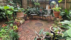 Um pátio circular de tijolos, com mesa e cadeiras de metal, rodeado por diversos vasos com plantas.