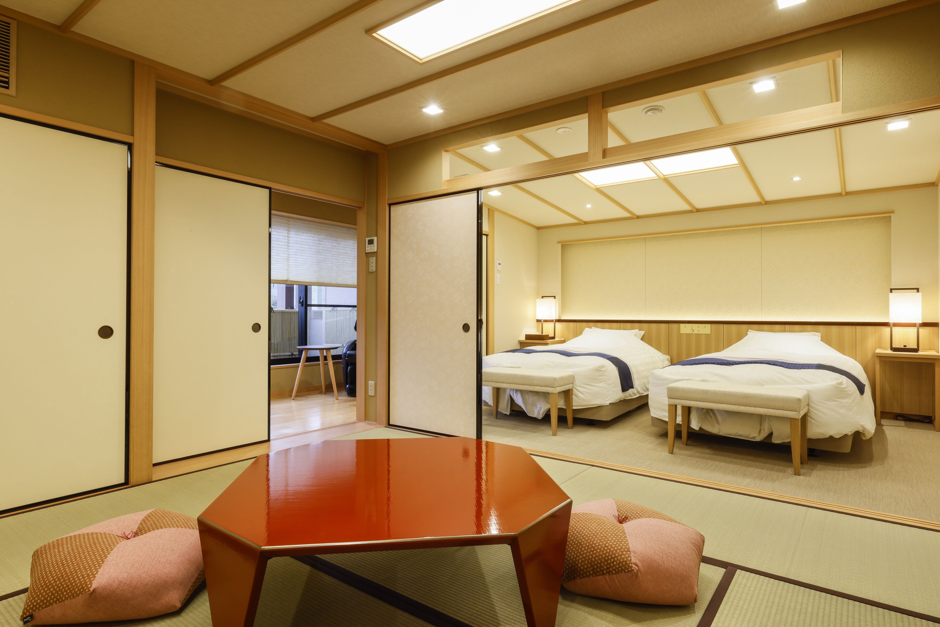 Une chambre d’hôtel de style japonais traditionnel, avec un sol recouvert de tatamis, une table en bois, deux lits et un grand miroir qui reflète la pièce.