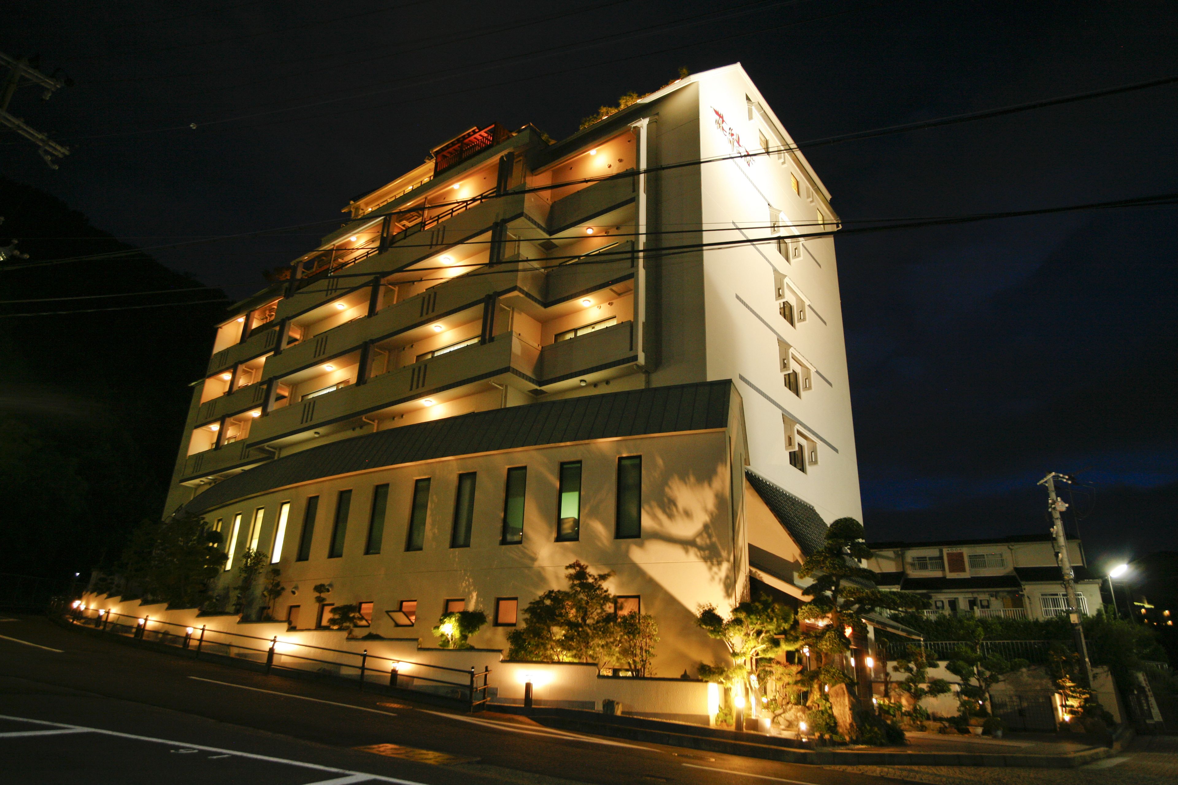 Un hotel de varios pisos con balcones, un exterior bien iluminado y un estacionamiento frente a él.
