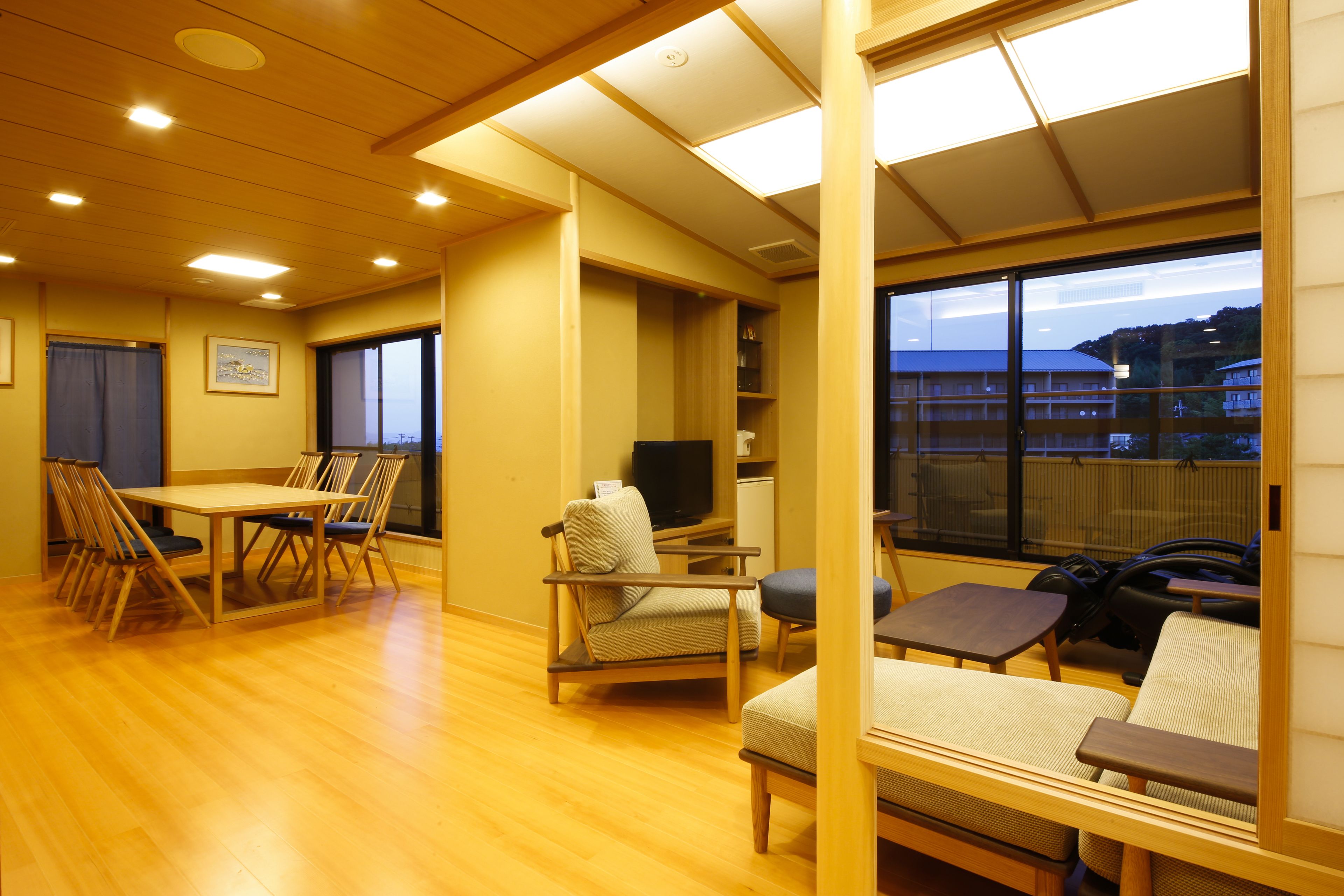 Une pièce spacieuse avec un plancher en bois, un coin repas comprenant des chaises et une table, un canapé, un téléviseur et une grande fenêtre donnant sur les bâtiments environnants.