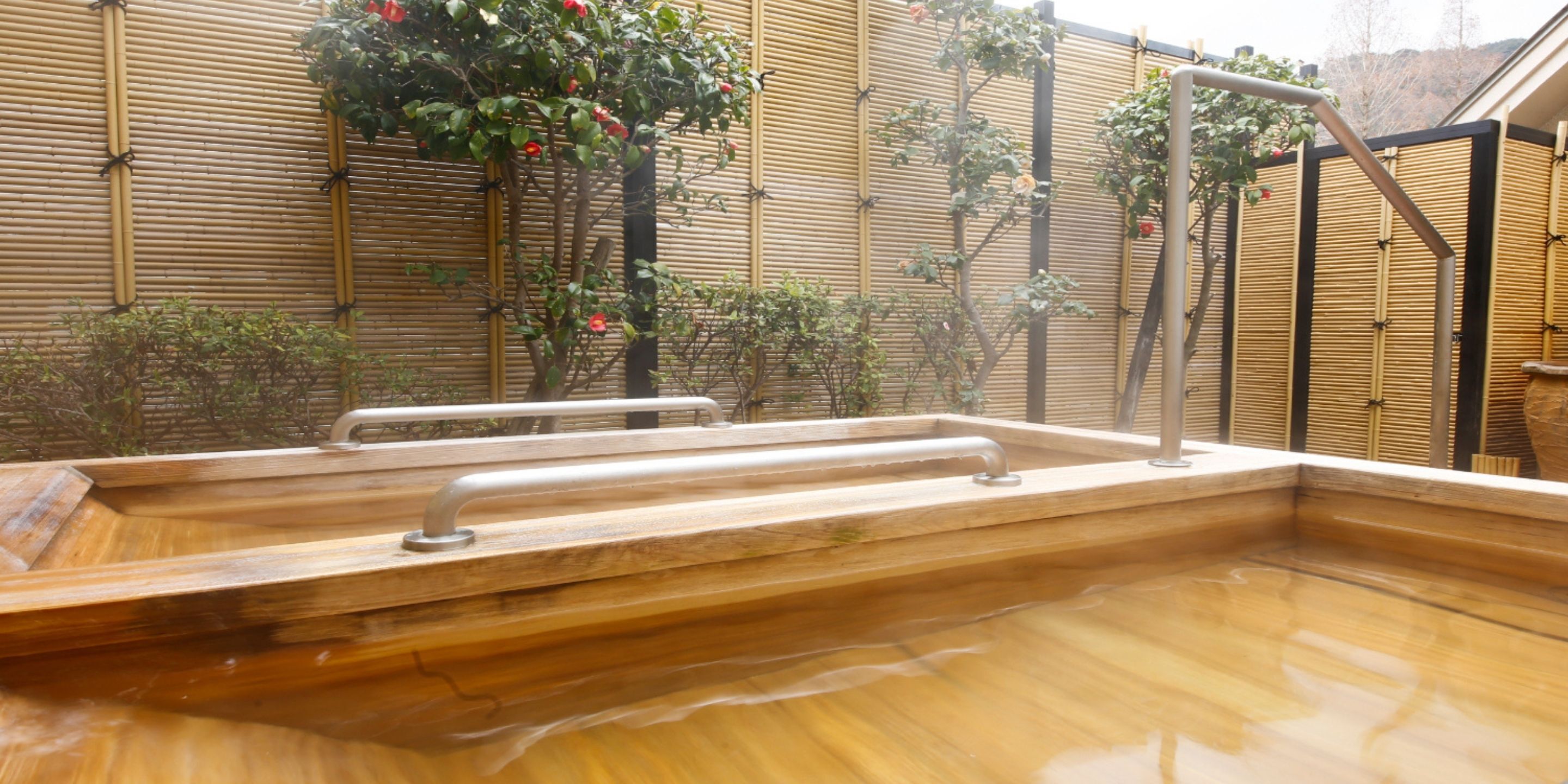 A wooden hot tub with metal railings, surrounded by bamboo walls and various plants.