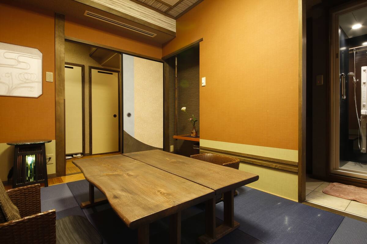 Una habitación con una mesa y sillas de madera, una mesita con lámpara, y una puerta corredera que da a otra habitación.