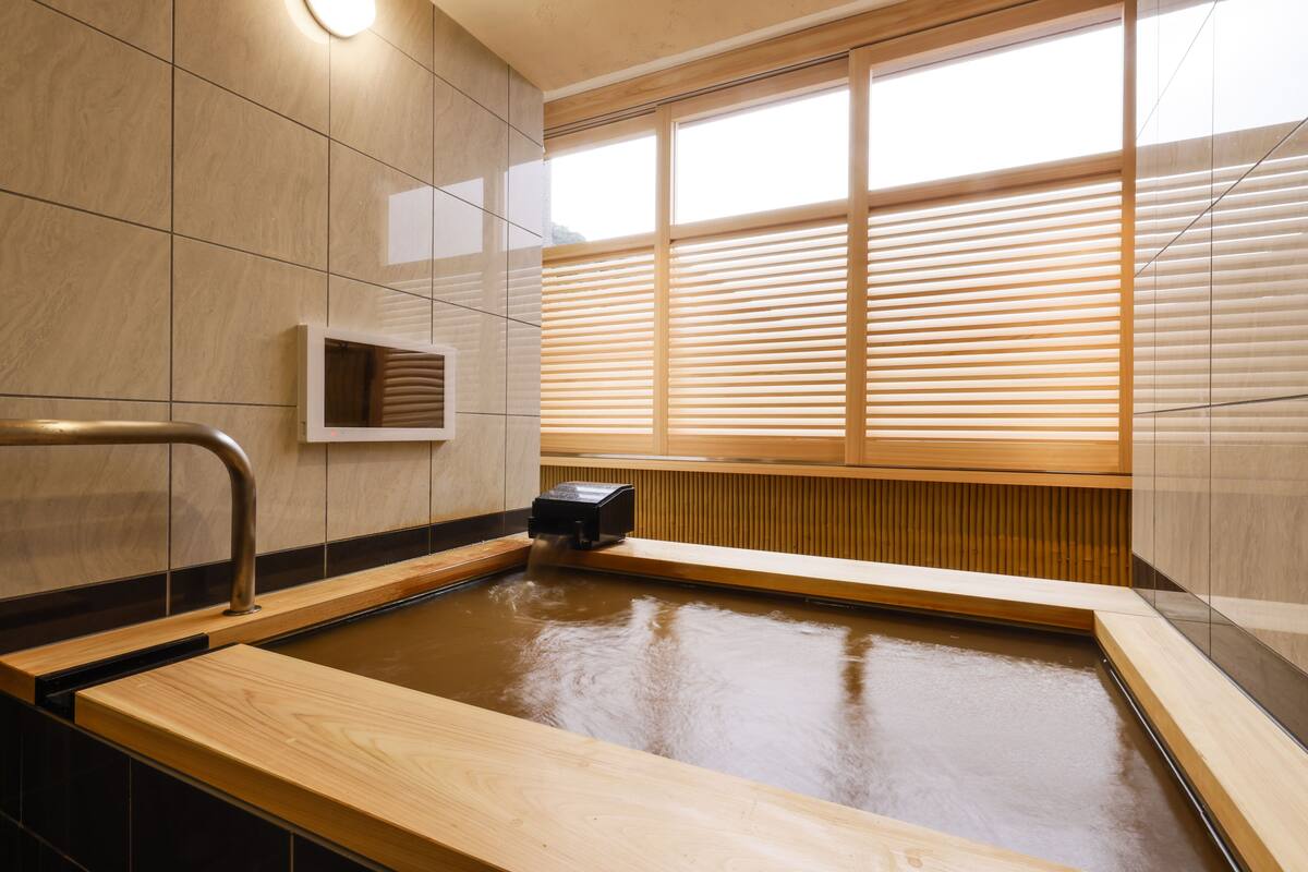 Un área de spa moderna con una bañera de madera, paredes de azulejos y una ventana con persianas.