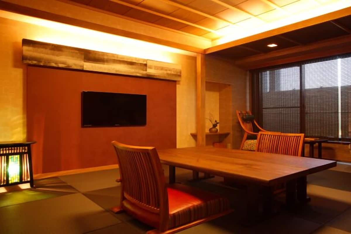 Une pièce comprenant une table à manger en bois, des chaises, un téléviseur à écran plat fixé au mur et une grande fenêtre avec des stores.