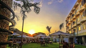 An outdoor dining area with tables and umbrellas, surrounded by palm trees and a building with balconies. - Hotel Centro Puerto Princesa, Palawan (Puerto Princesa)