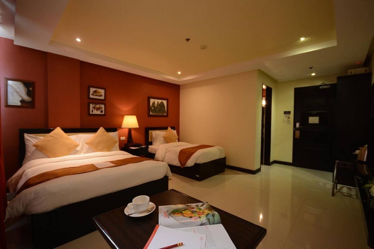 A hotel room with two beds, a desk with a cup and papers, and a door to the right.