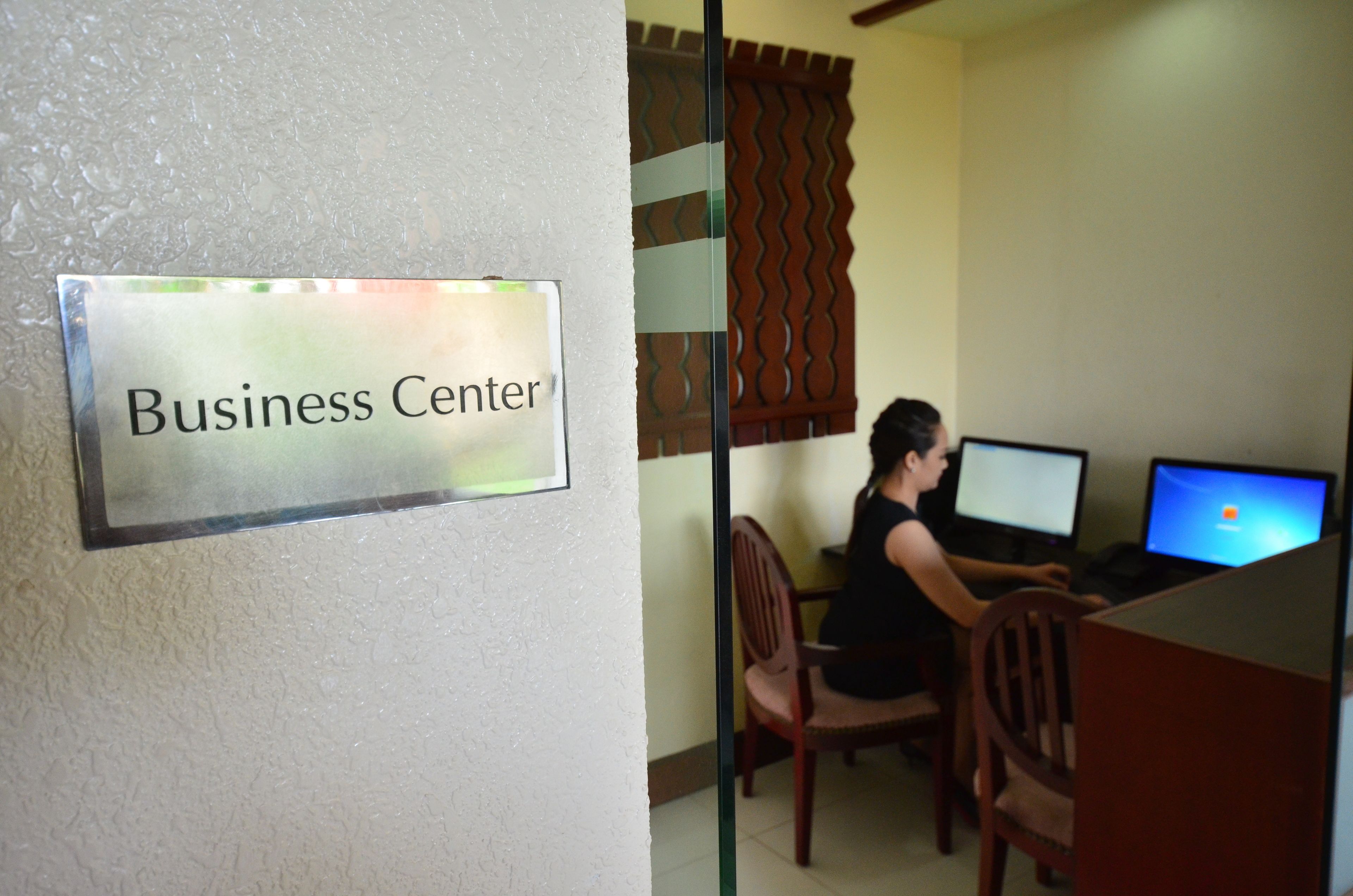 A business center with a sign, a person working at a desk with two computers, and wooden chairs.