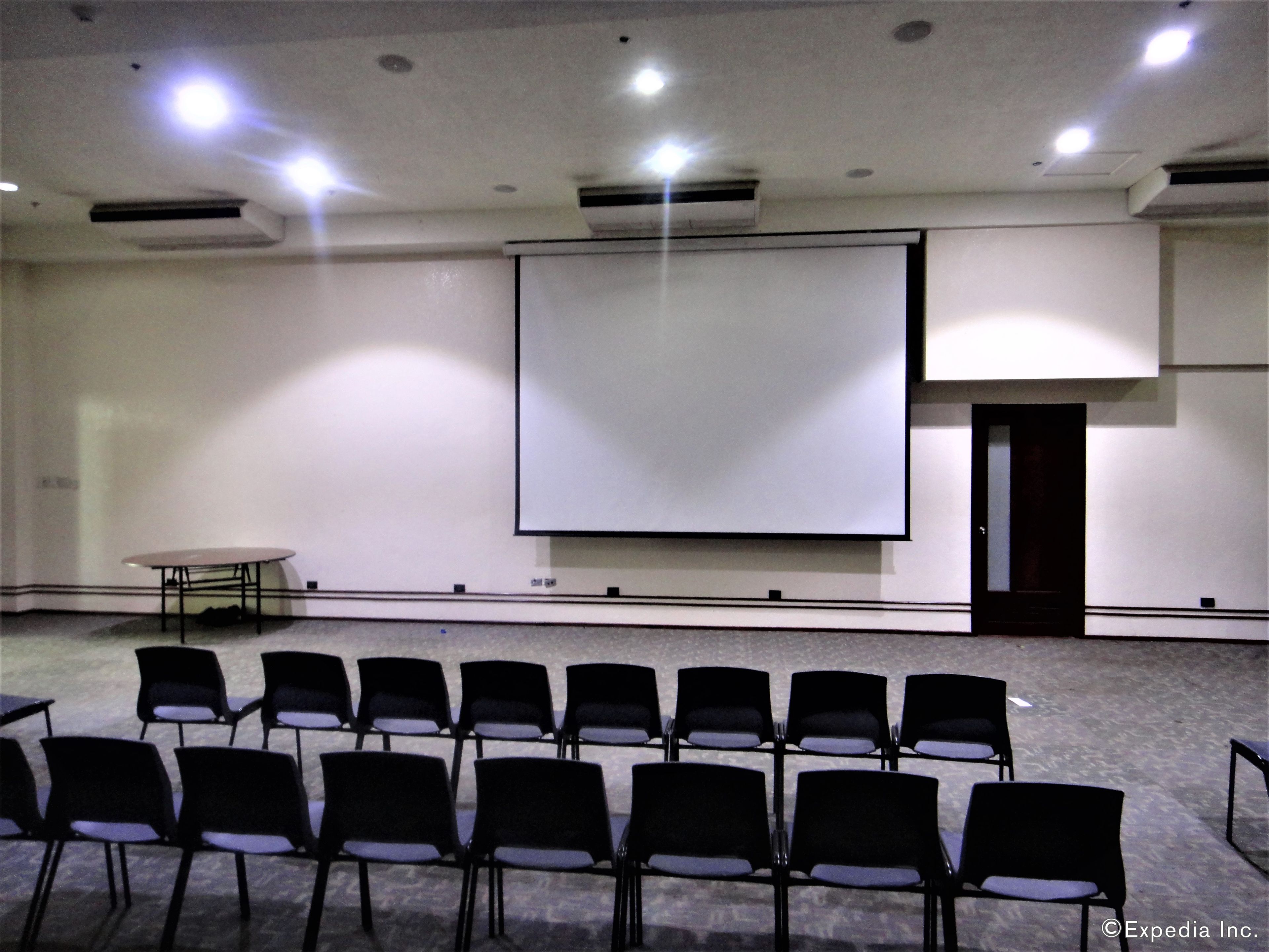 Un salón de conferencias con una pantalla de proyección grande, filas de sillas y una mesa.