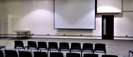 Un salón de conferencias con una pantalla de proyección grande, filas de sillas y una mesa.