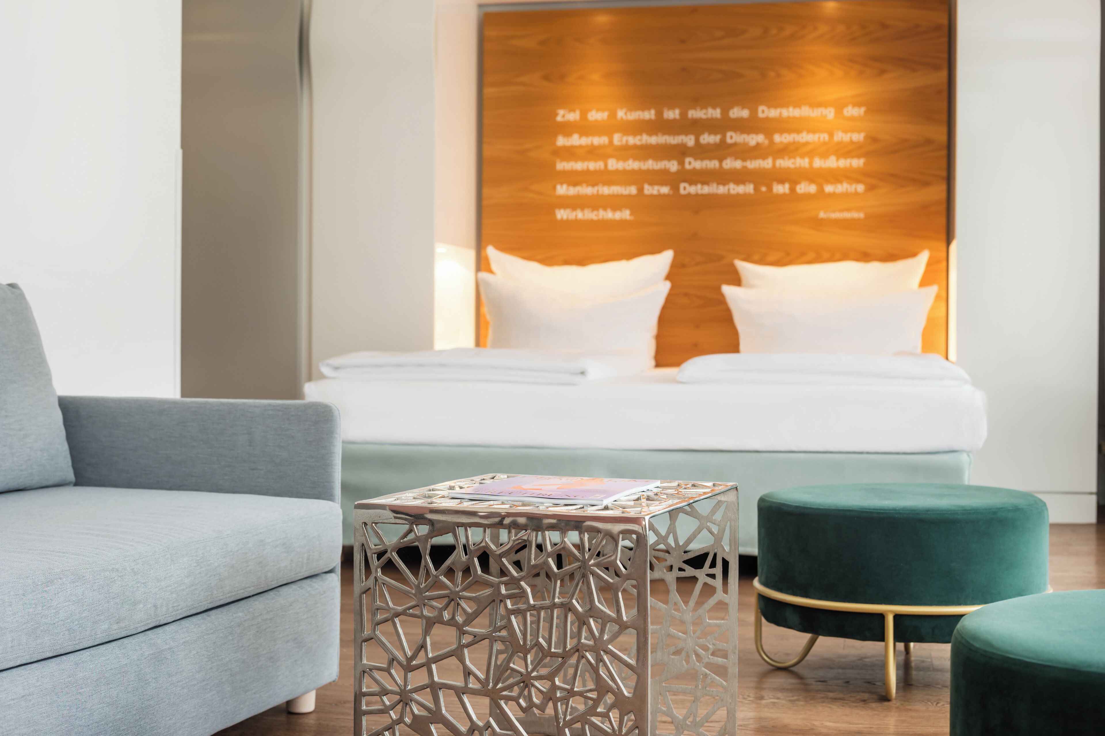 A modern hotel room with a bed, a grey sofa, a decorative table, and a green ottoman.
