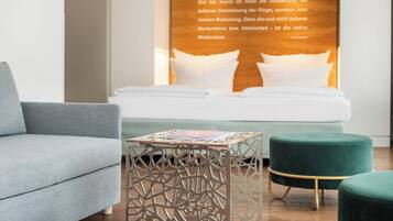 A modern hotel room with a bed, a grey sofa, a decorative table, and a green ottoman.