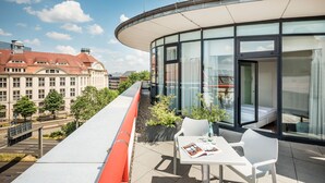 A modern building with a curved glass facade, a rooftop terrace with a white table and chairs, and a view of the cityscape in the background. - Art Hotel City Leipzig (Leipzig)
