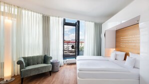 A modern hotel room with a large bed, a sofa, a small table, and a view of the sky through a large window. - Art Hotel City Leipzig (Leipzig)