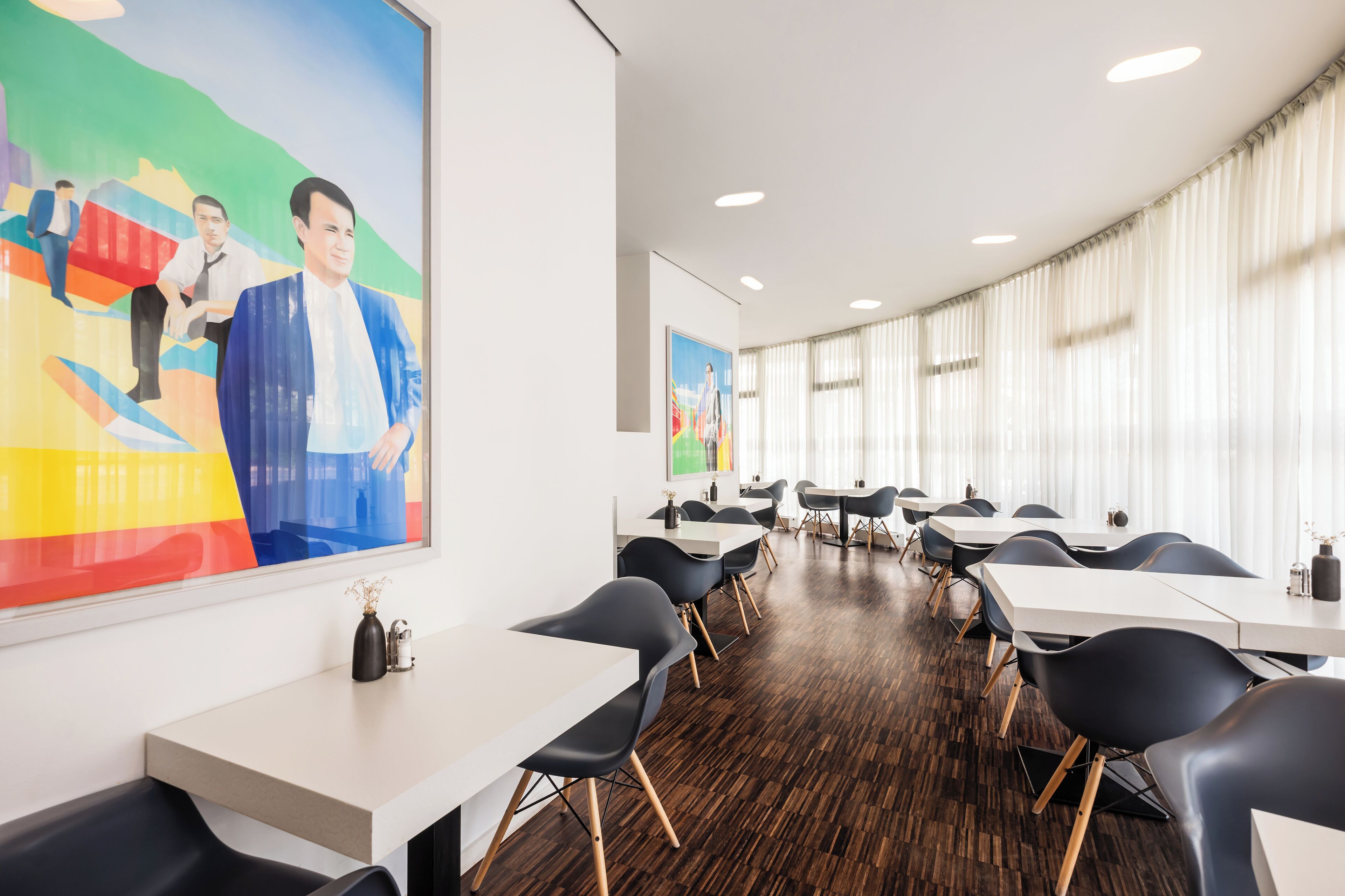 Une salle de conférence moderne, avec des tables blanches et des chaises noires, de grandes fenêtres ornées de rideaux légers et une peinture abstraite aux couleurs vives accrochée au mur.