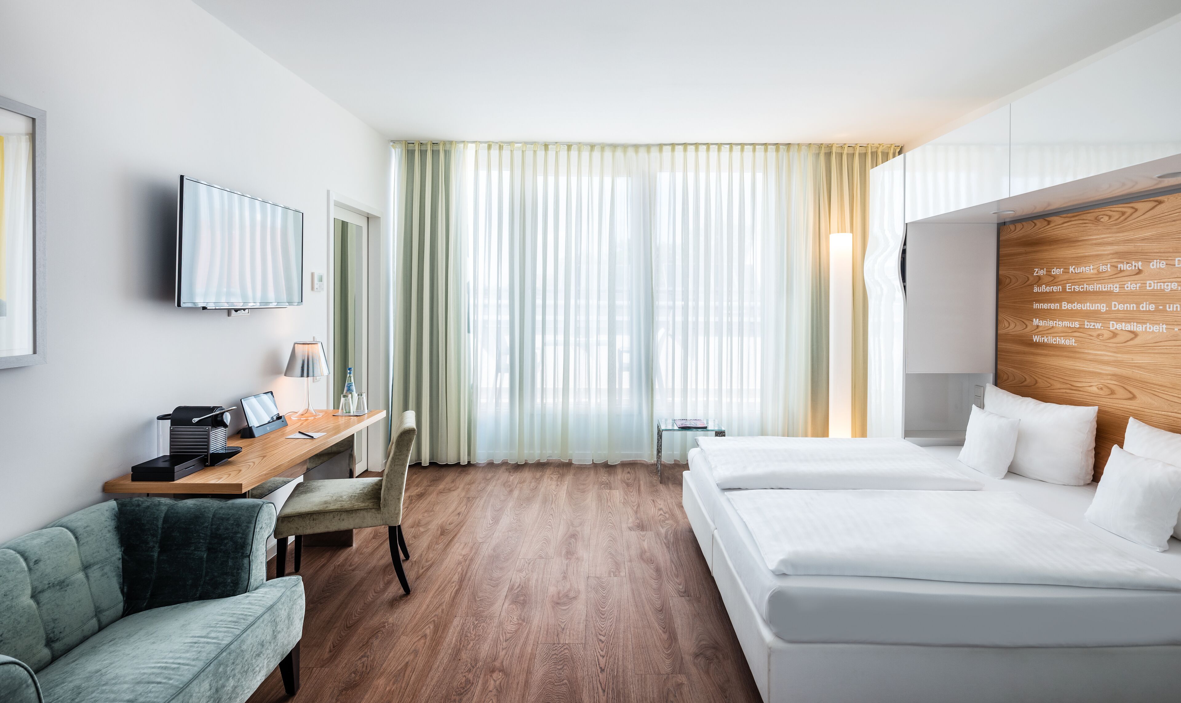 A modern hotel room with a wooden floor, a desk with a laptop, a chair, a bed with white linens, and a large window with sheer curtains.