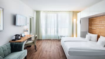 A modern hotel room with a wooden floor, a desk with a laptop, a chair, a bed with white linens, and a large window with sheer curtains.