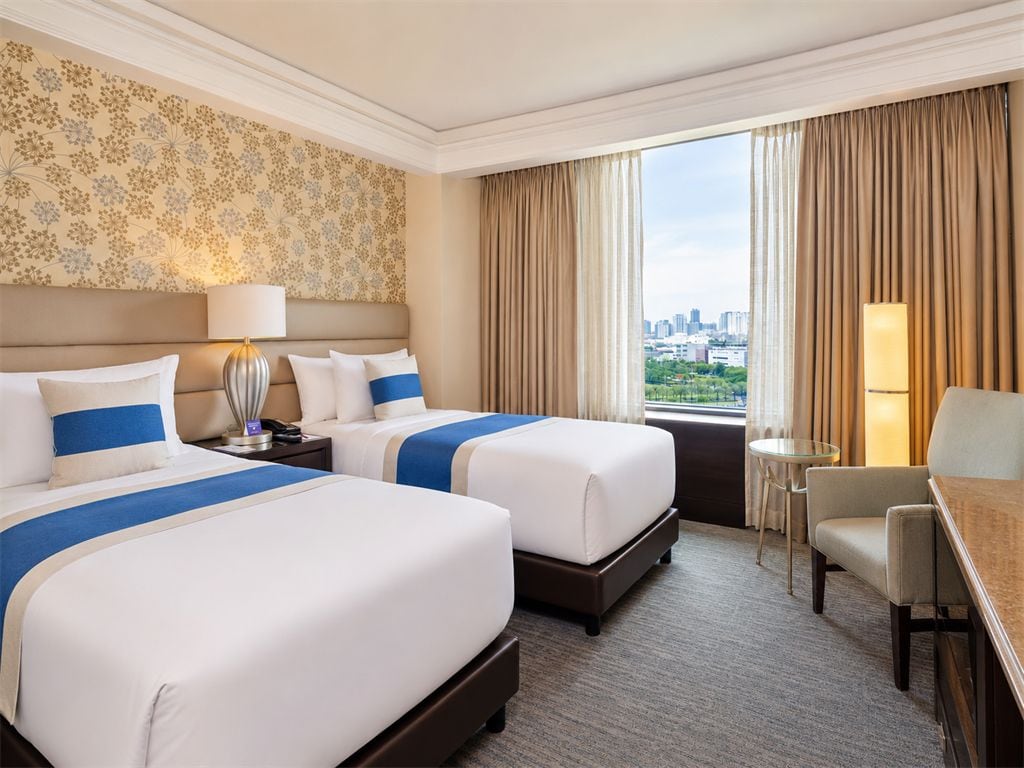 A hotel room with two beds, a desk, a chair, and a view of the city.