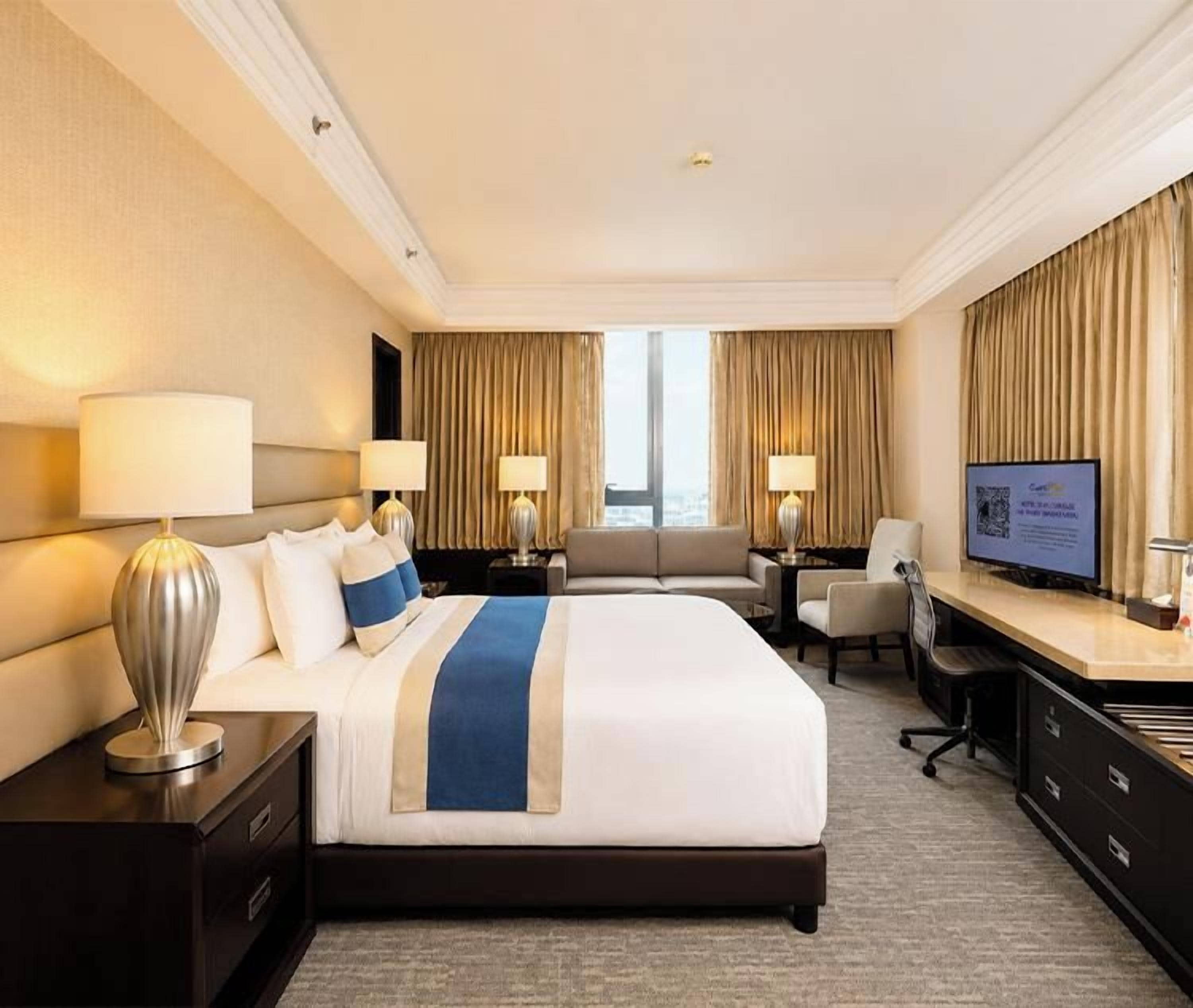 A hotel room with a large bed, two bedside tables with lamps, a desk with a chair, a television, and a window with curtains.