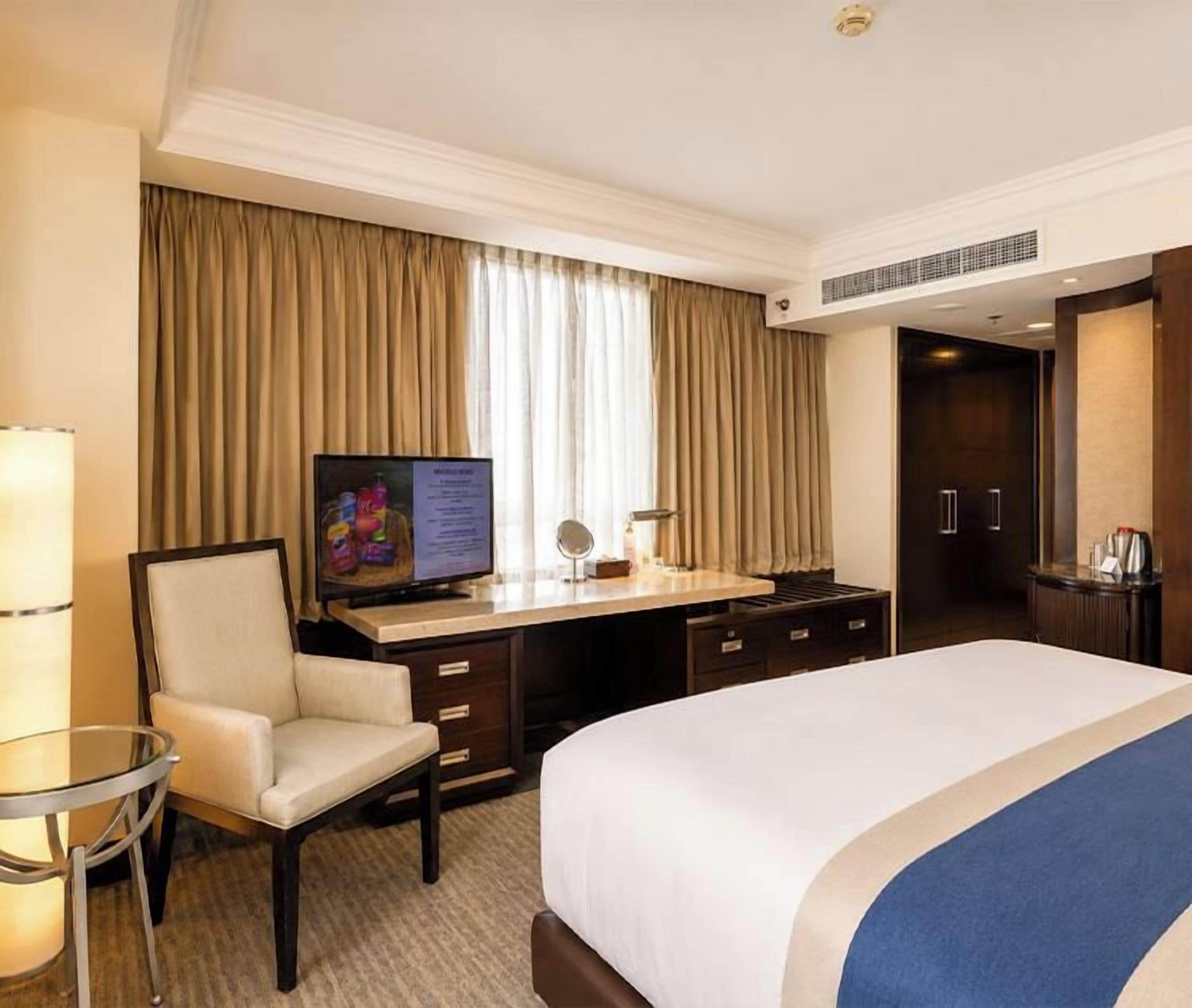 Habitación de hotel con cama, escritorio, silla, televisor y ventana con cortinas.
