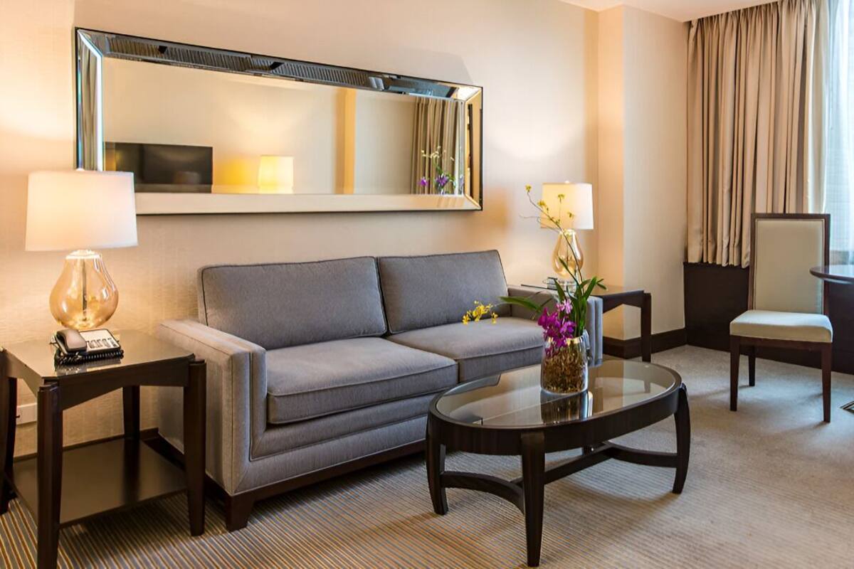A hotel room with a sofa, a glass coffee table, a chair, a lamp, and a mirror.