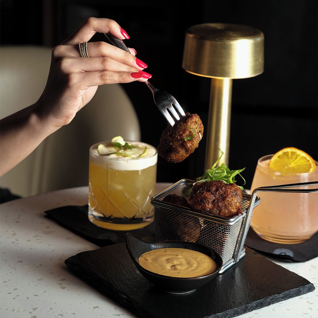 A hand holding a fork with a food item, a glass of beer, a cocktail, and a small bowl of sauce on a table.