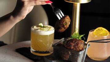 A hand holding a fork with a food item, a glass of beer, a cocktail, and a small bowl of sauce on a table.