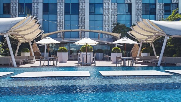 A modern hotel pool area with a clear view of the building facade, outdoor seating, and a central water feature.
