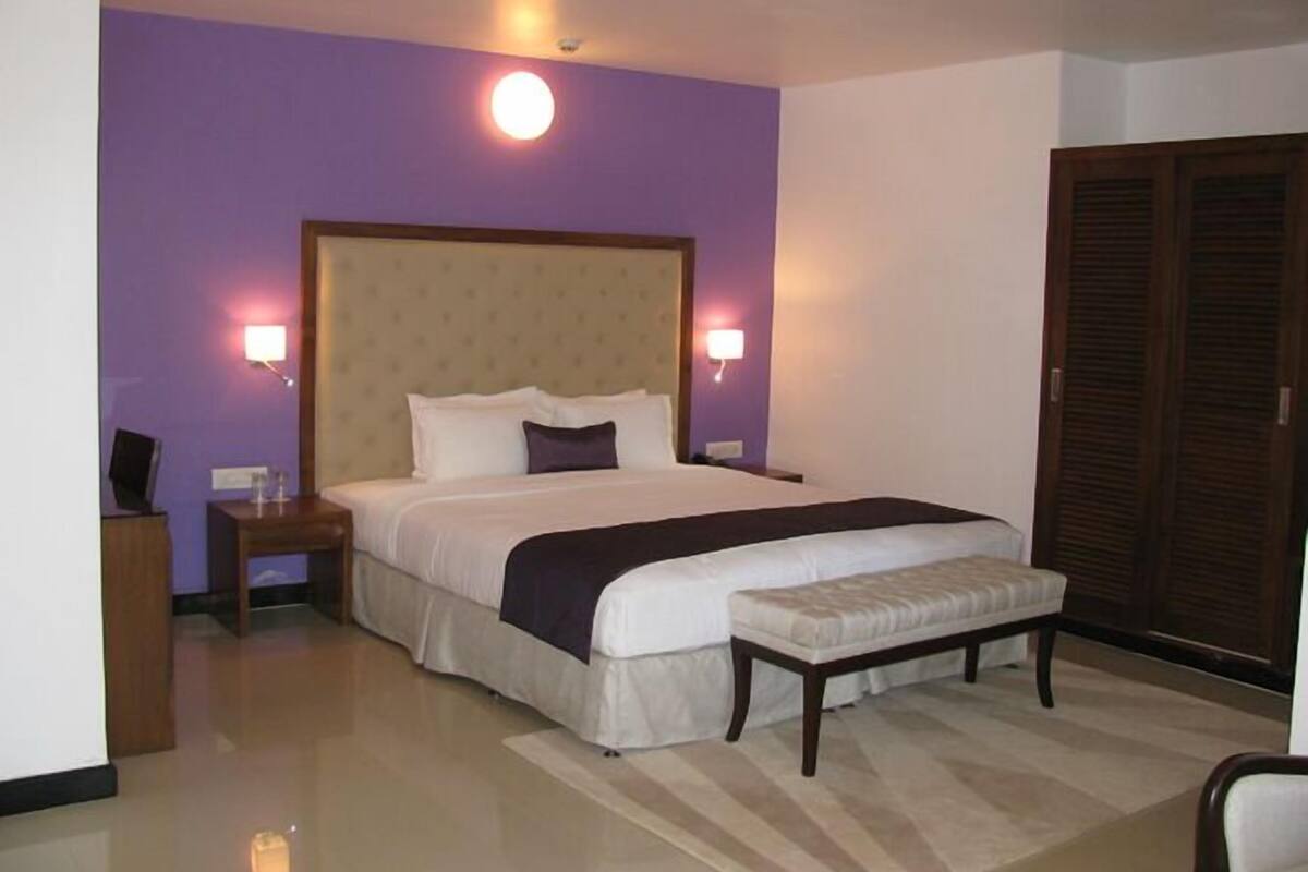Una habitación de hotel con una cama grande, dos lámparas de noche, un banco de madera, una mesita de noche y un armario.