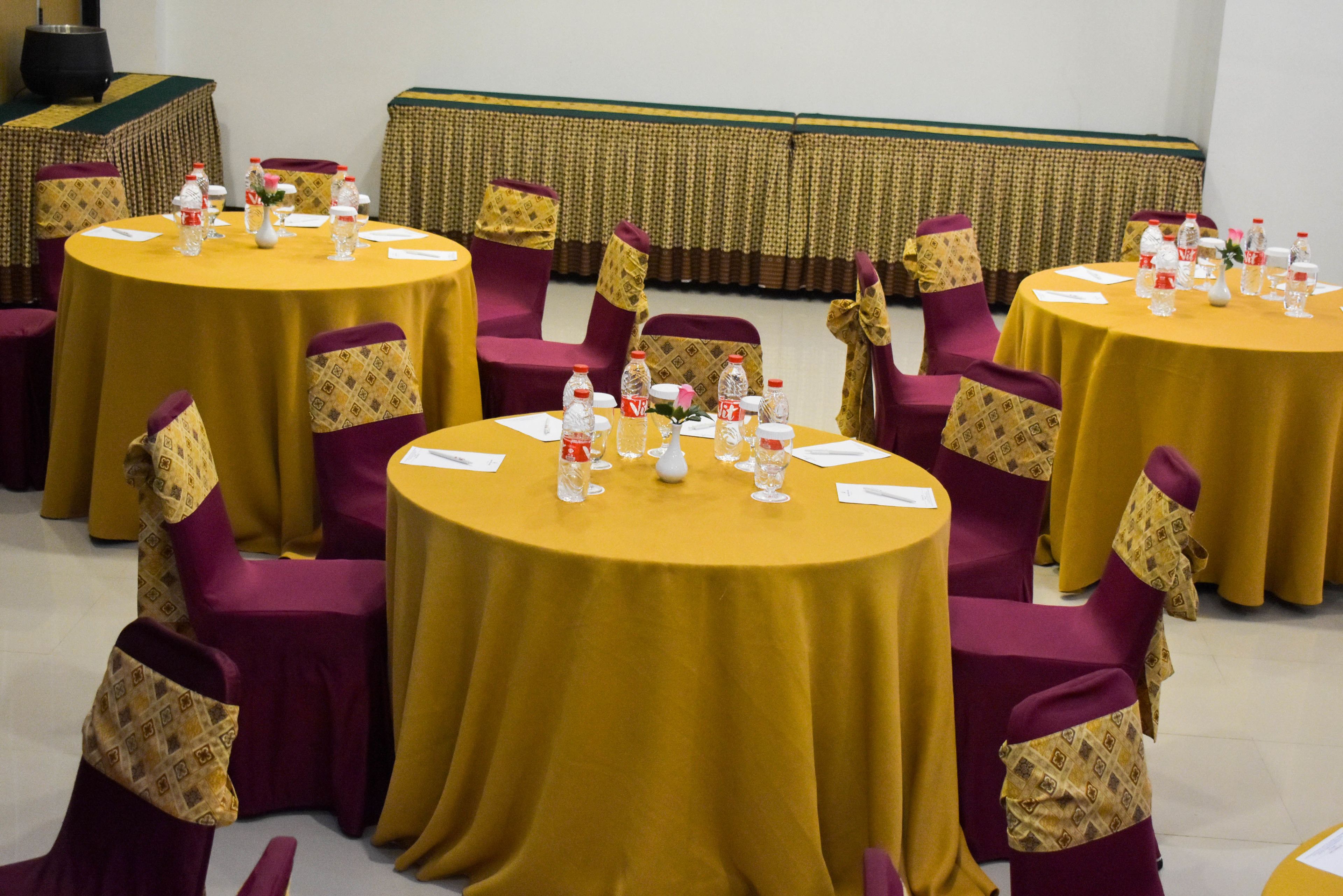 Ein Konferenzraum mit mehreren runden Tischen, jeder mit einer gelben Tischdecke und umgeben von violetten Stühlen mit gemusterten Kissen.