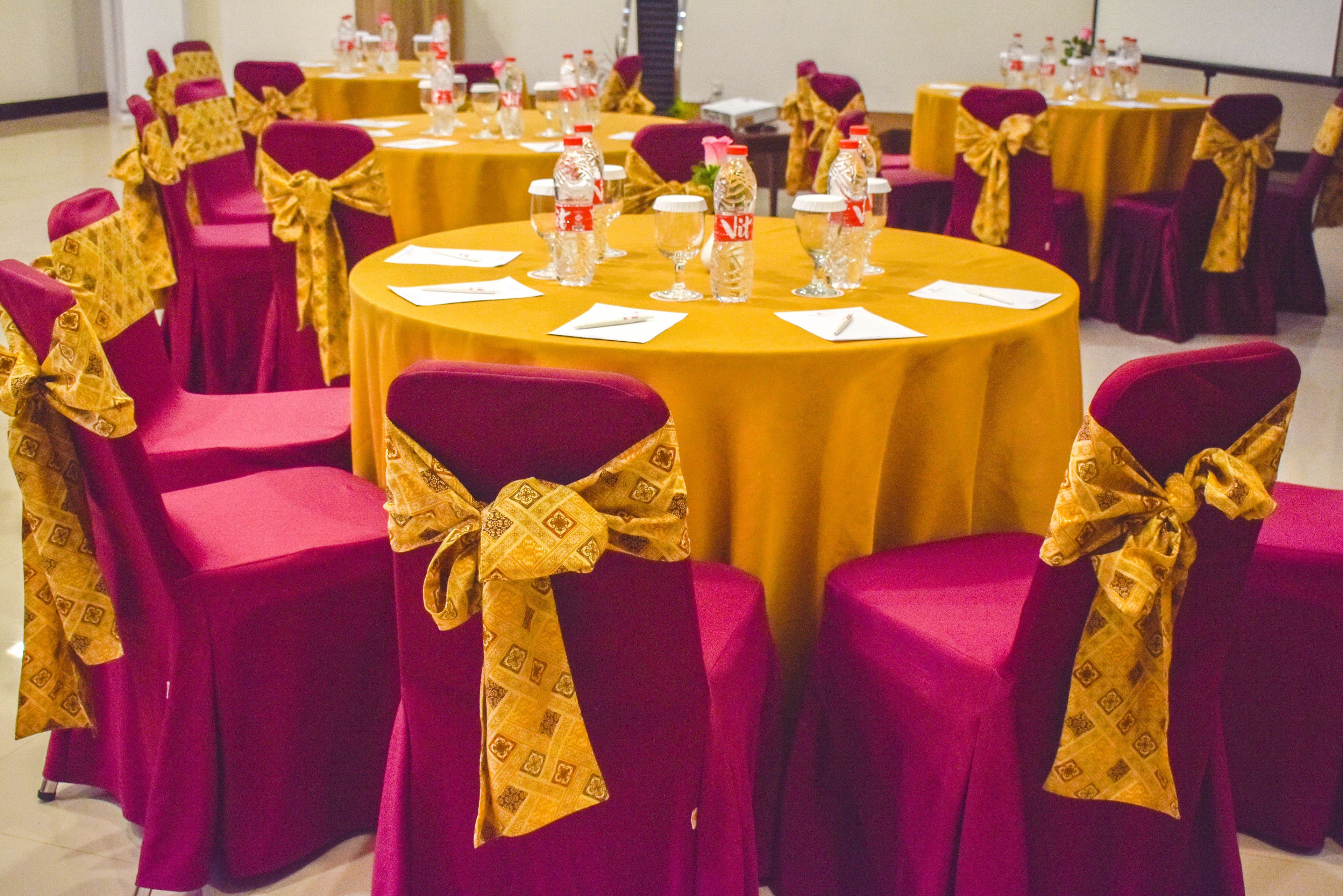 Een vergaderzaal met ronde tafels, elk omringd door stoelen met gouden linten. Op de tafels staan waterflesjes en glazen.