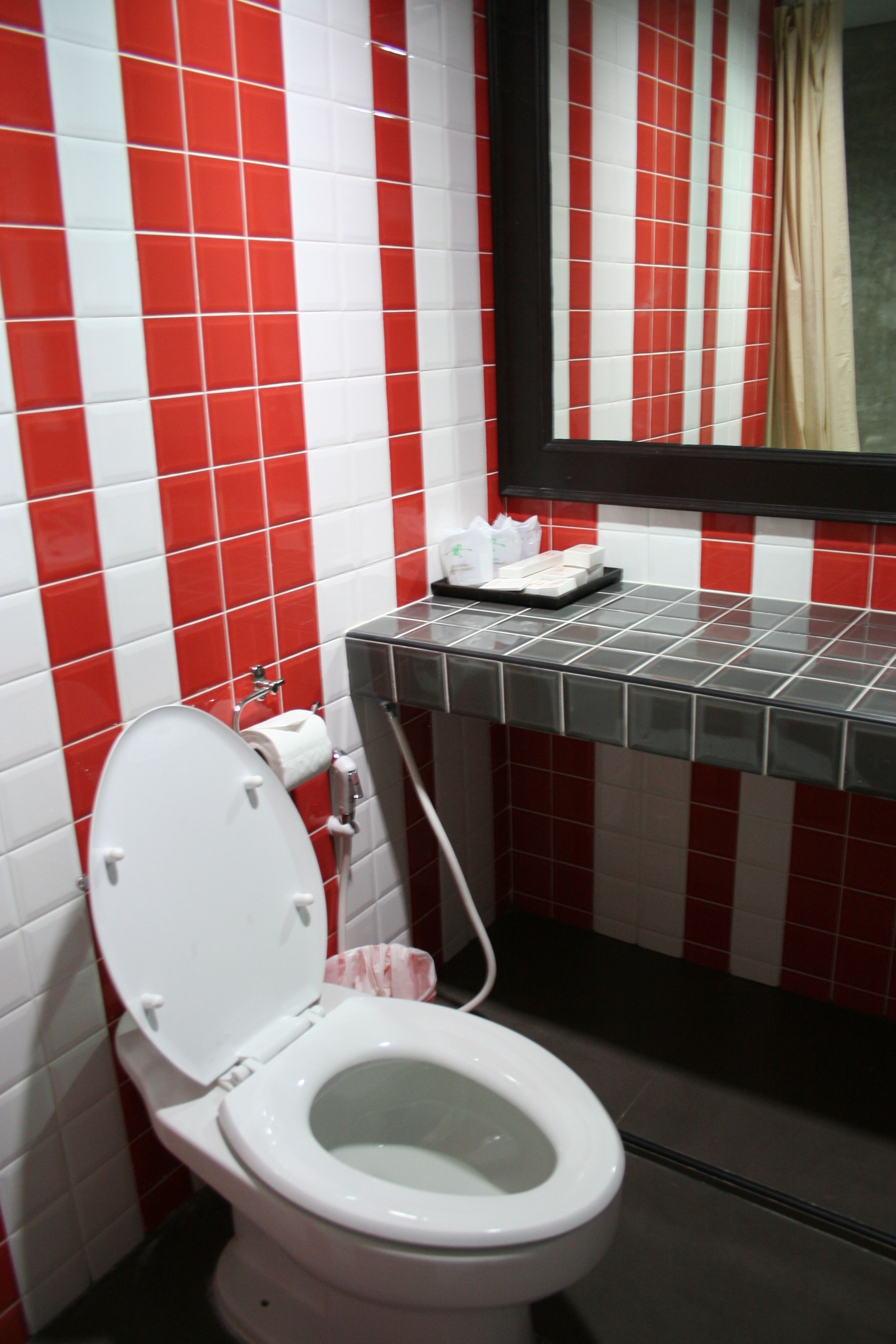 Un baño con paredes cubiertas de azulejos blancos y rojos, un inodoro blanco y un espejo encima.