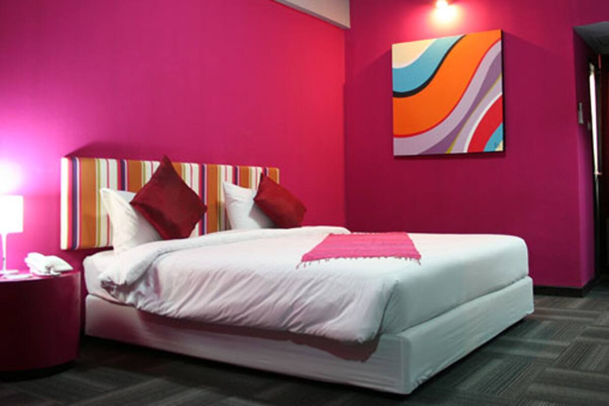 A hotel room with a bed, a bedside table with a lamp, and a colorful abstract painting on the wall.