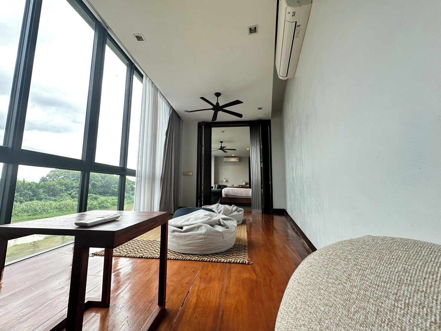 A modern hotel room with large windows, a wooden floor, a ceiling fan, and a bed with a nightstand.